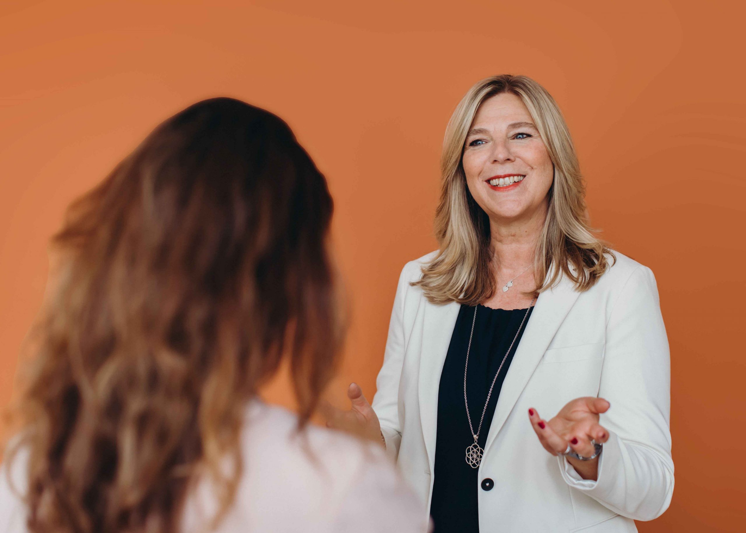 Christina Becker in einem professionellen Gespräch mit einer Person im Vordergrund. Sie trägt einen weißen Blazer über einem dunklen Oberteil, spricht gestikulierend und lächelt. Der Hintergrund ist in einem warmen Orangeton gehalten.